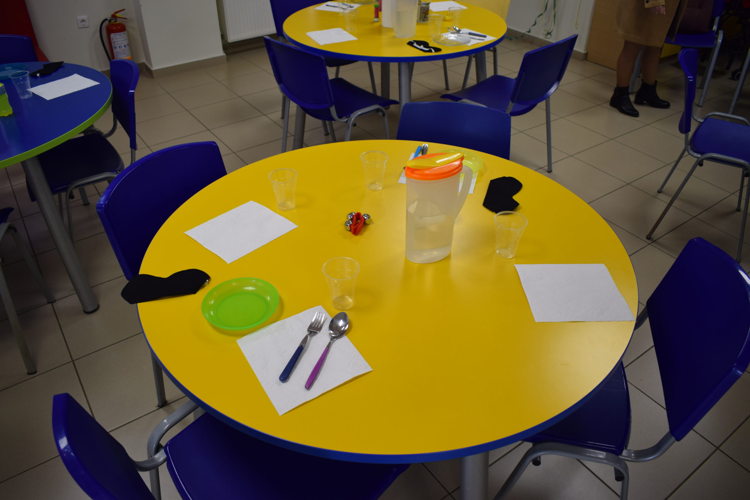 Στη φωτογραφία φαίνεται ένα κίτρινο τραπέζι στην τραπεζαρία της Συζωής που έρχεται σε έντονη οπτική αντίθεση με τις μπλε καρέκλες, το πράσινο πιάτο και την πορτοκαλί κανάτα για νερό.